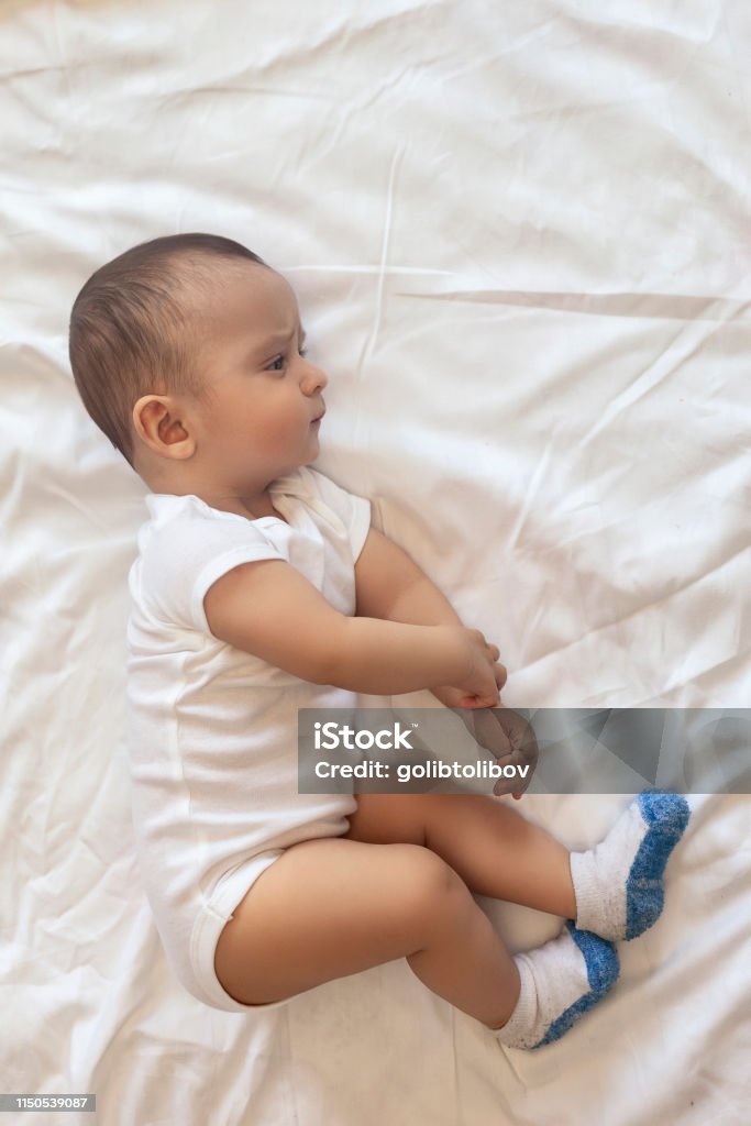 6-8 month old baby boy lying playfully in bed 6-8-month-old baby boy lying playfully in bed. Charming 6-7 month little baby in white bodysuit. Baby boy in white bedding. Free space 0-11 Months Stock Photo