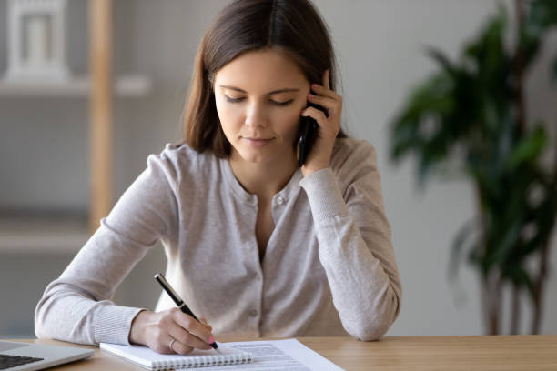 zajęta atrakcyjna kobieta siedząca przy stole rozmawiając przez telefon - smart phone writing assistance business zdjęcia i obrazy z banku zdjęć