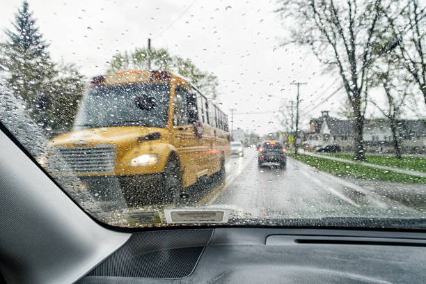 A large yellow school bus approaching from the opposite direction during a pouring rain storm downpour is passing to the left of this stationary vehicle. Car driver personal perspective - car point of view looking through windshield.