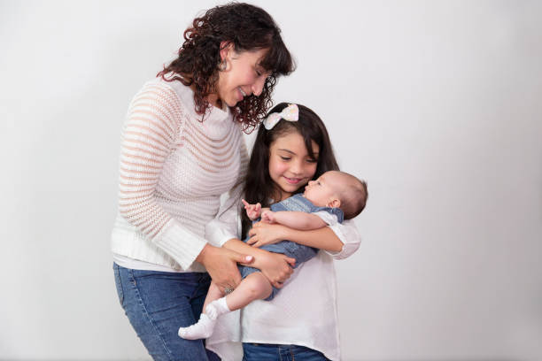 7 year old girl with her little sister in her arms smiling while her mother helps hold her with love - latin family - crimped imagens e fotografias de stock