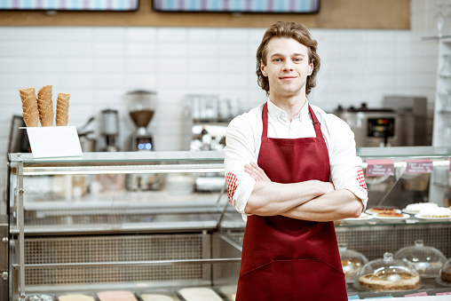 Portrait of a handsome salesman or waiter in red apron standing in the pastry cafe with shop-front on the background