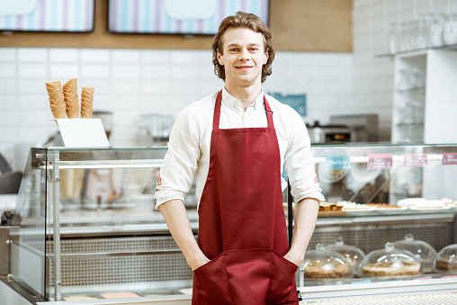 Portrait of a handsome salesman or waiter in red apron standing in the pastry cafe with shop-front on the background