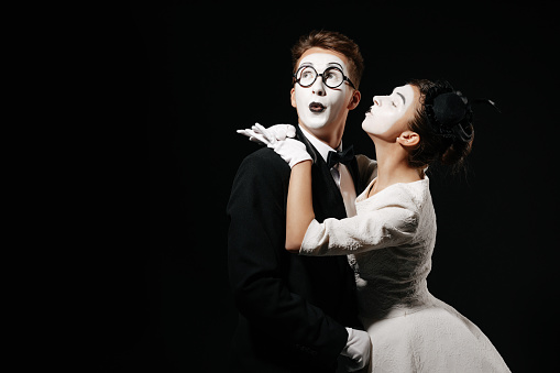 portrait of couple mime on black background. woman in white dress kissing man in tuxedo and glasses