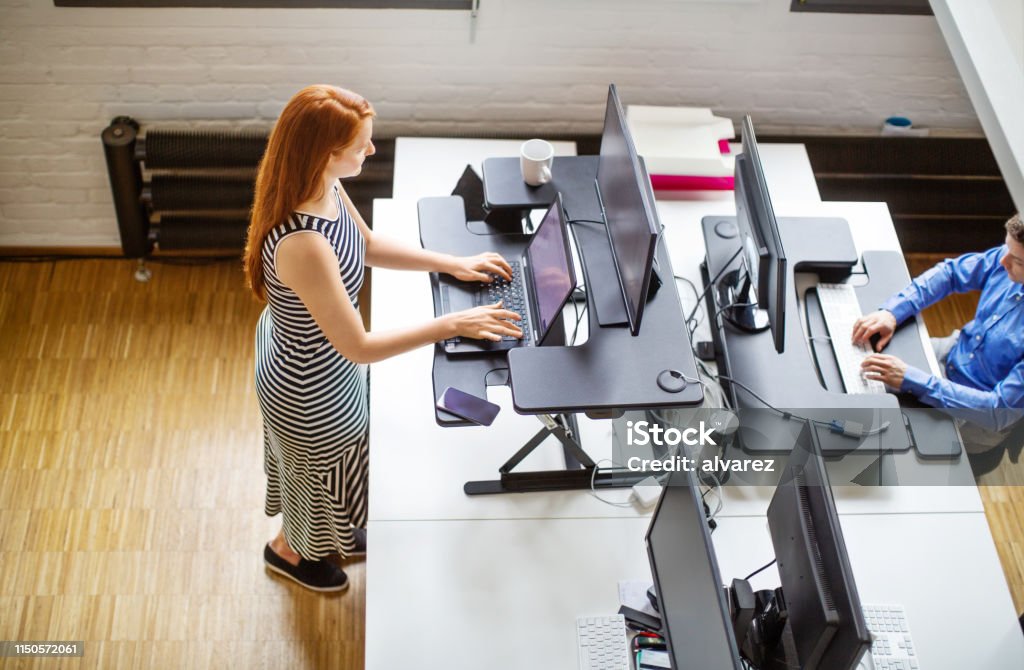 Woman working at ergonomic standing desk Top view of woman employee working on laptop computer at ergonomic standing desk. Female professional working at her desk with male colleague working around the table. Standing Desk Stock Photo