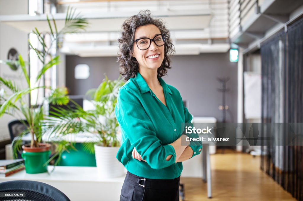 Portrait of confident mature businesswoman in office Portrait of happy mature businesswoman standing with her arms crossed. Mid adult female looking at camera and smiling while standing in office. Women Stock Photo