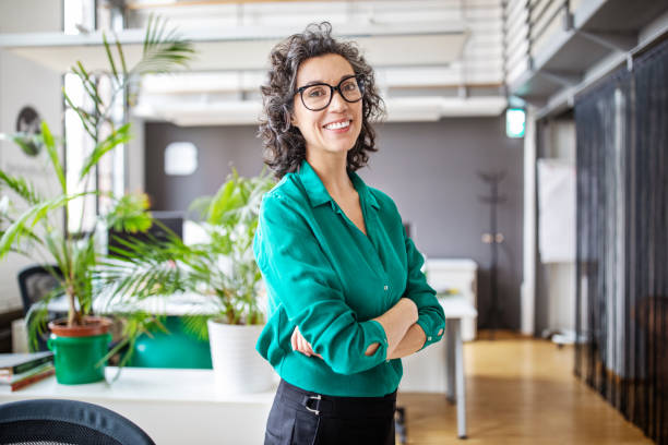 Portrait of confident mature businesswoman in office Portrait of happy mature businesswoman standing with her arms crossed. Mid adult female looking at camera and smiling while standing in office. business casual stock pictures, royalty-free photos & images
