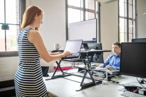 Business woman working on laptop computer at ergonomic standing desk. Female professional working at her desk with male colleague working at the back.