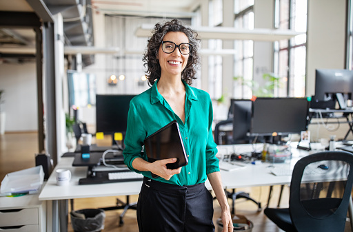 Portrait of mature businesswoman in casuals standing at her desk with digital tablet. Woman entrepreneur in office looking at camera and smiling.