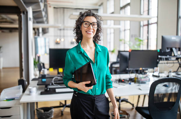 Portrait of mature businesswoman in casuals Portrait of mature businesswoman in casuals standing at her desk with digital tablet. Woman entrepreneur in office looking at camera and smiling. three quarter length photos stock pictures, royalty-free photos & images