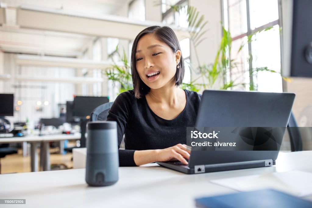 Female professional using virtual assistant at desk Asian businesswoman talking to virtual assistant at her desk. Female professional working on laptop and talking into a speaker. Artificial Intelligence Stock Photo