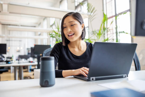 Female professional using virtual assistant at desk Asian businesswoman talking to virtual assistant at her desk. Female professional working on laptop and talking into a speaker. bluetooth stock pictures, royalty-free photos & images