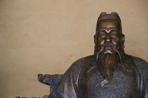 Statue of sad tired old Chinese man with big eyes, a whispy beard, ornately embroidered shirt and a hat that looks like a plastic block brick. Made from a golden brown metal (likely bronze)