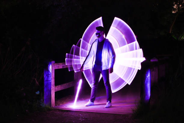 Portrait of a man in a surrealistic setting done at lightpainting with light effects Boy with a mask and a lightsaber and virtual wings, standing on a wooden bridge at night lightpainting stock pictures, royalty-free photos & images