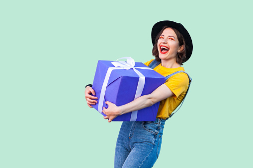 Satisfied happy beautiful young girl in hipster wear in denim overalls and black hat standing and holding big heavy gift box with toothy smile and laughing, open mouth.