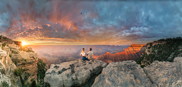 This is a panoramic photograph of a man and a woman sitting on the edge of rim, talking about future and watching the Grand Canyon sunset while bird in the sky