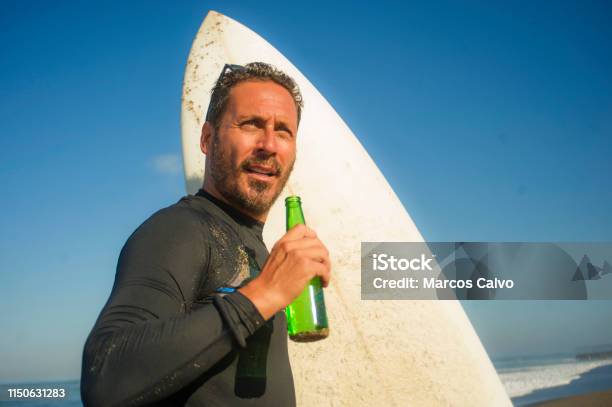 Closeup Portrait Of Handsome And Attractive Surfer Man In Neoprene Swimsuit Holding Surf Board Posing Cool And Happy After Surfing Enjoying Summer Water Sport And Holidays Drinking Beer Bottle Stock Photo - Download Image Now