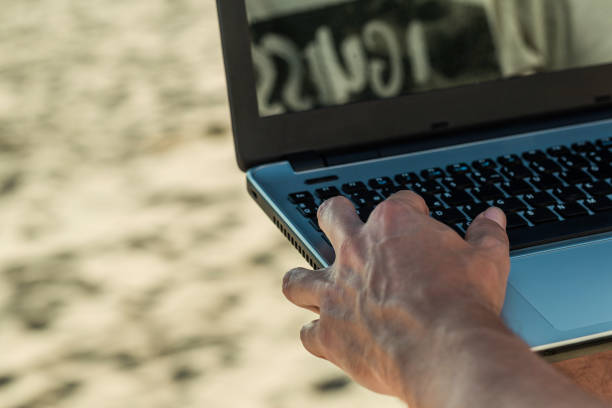 Close up hand typing laptop with blank screen on wooden table over sand background. Freelance working at anywhere and using social media communication concept