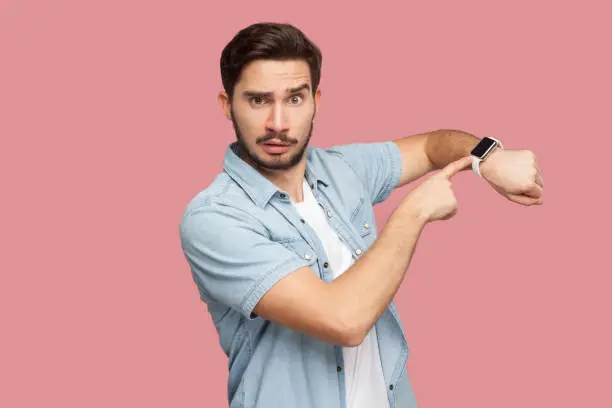 Photo of Time is out. Portrait of serious handsome bearded young man in blue casual style shirt standing and looking at camera, pointing on his smart watch.