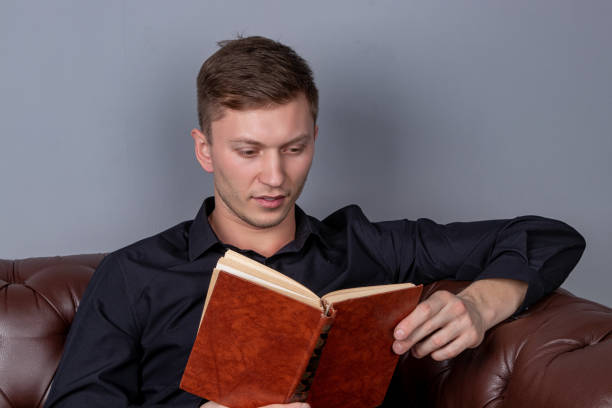 istock Young man is sitting on a leather sofa and reading a book 1150684743