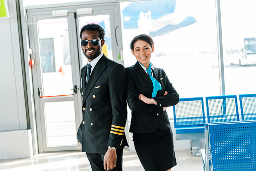 smiling african american pilot in sunglasses and stewardess with crossed arms standing together in airport