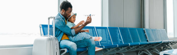panoramic shot of african american father and son sitting with suitcase in airport and playing with toy plane panoramic shot of african american father and son sitting with suitcase in airport and playing with toy plane toy airplane stock pictures, royalty-free photos & images
