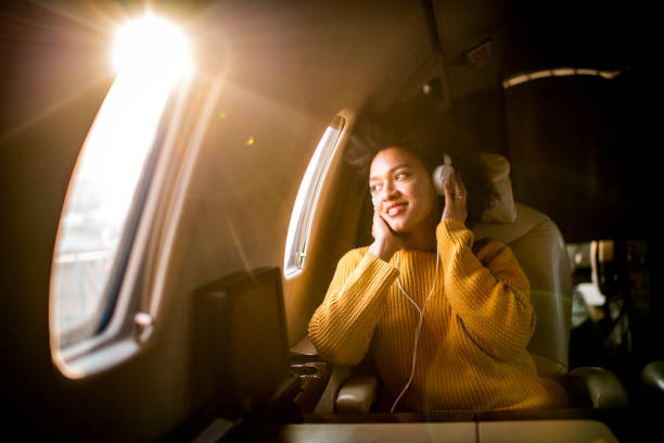 joven mujer de moda sentada en un avión privado y mirando a través de una ventana mientras escucha música a través de auriculares - status symbol audio fotografías e imágenes de stock