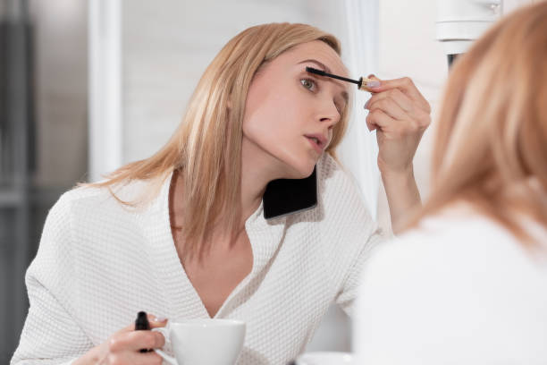 attraente donna bionda in accappatoio bianco sta in bagno per specchio. applica il trucco, parla al telefono cellulare e beve caffè. tasse mattutine, in qualsiasi momento, facendo molta azione allo stesso tempo. - bathrobe women cup towel foto e immagini stock