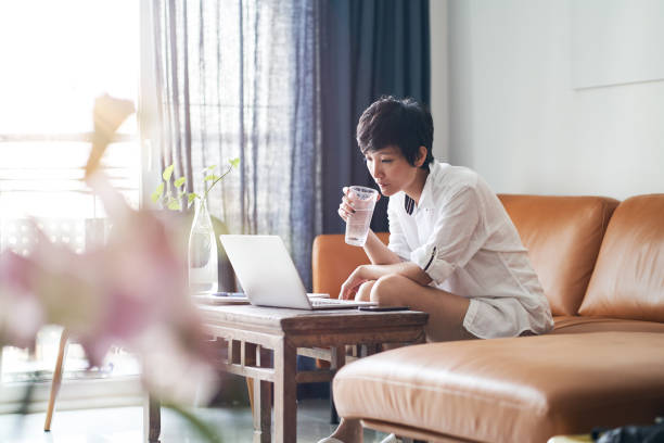 bellezza asiatica per adulti seduta sul divano e lavorando su laptop da casa - house home interior water glass foto e immagini stock