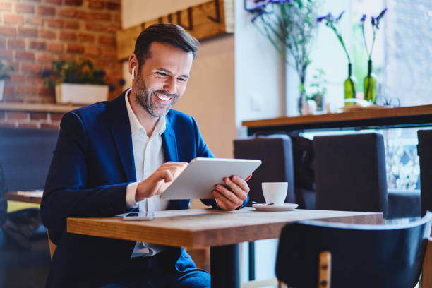 businessman sitting in cafe using tablet and talking through wireless headphones - business business person ceo coffee imagens e fotografias de stock