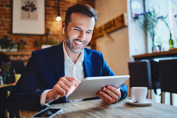 geschäftsmann sitzt im café und schaut sich tablet beim telefonieren über drahtlose kopfhörer an - restaurant sitting adult beauty stock-fotos und bilder