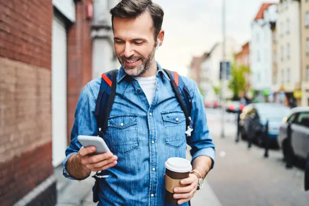 Photo of Man using phone with wireless headphones walking in the city and drinking coffee