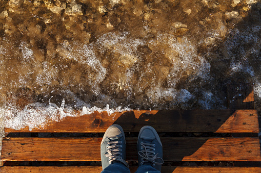 The tourist stopped on a Wooden bridge to look at the water and the clear waves of the river on the rocky bottom. Selfie shoes, top view