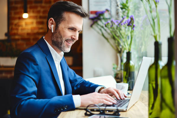 hombre de negocios feliz hablando a través de auriculares inalámbricos mientras trabaja en el ordenador portátil en café - hot drink audio fotografías e imágenes de stock