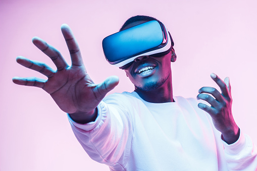 Hombre afroamericano en gafas VR, jugando videojuegos con cascos de realidad virtual, tratando de tocar algo con la mano photo
