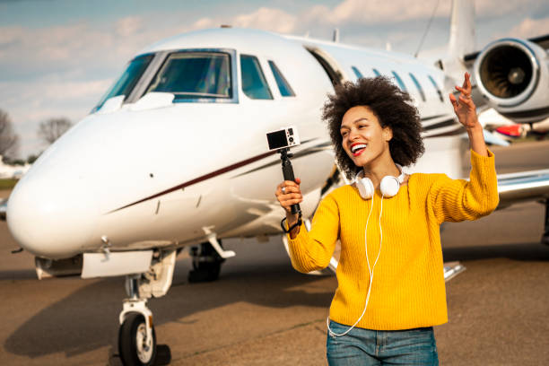 joven vlogger hembra haciendo un espectáculo con su cámara montada en un palo selfie junto a un jet privado estacionado en una pista de aterrizaje del aeropuerto - status symbol audio fotografías e imágenes de stock