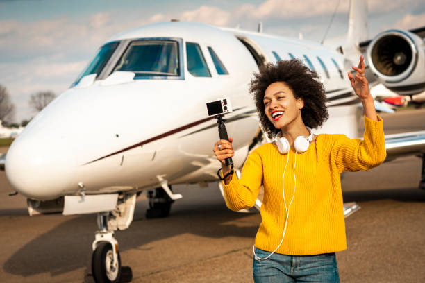 jeune vlogger femelle faisant un spectacle avec son appareil-photo monté sur un bâton de selfie à côté d’un jet privé stationné sur une piste d’aéroport - status symbol audio photos et images de collection