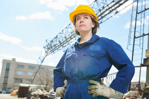 Waist up portrait of empowered woman working at construction site standing against sky with tower crane in background, copy space