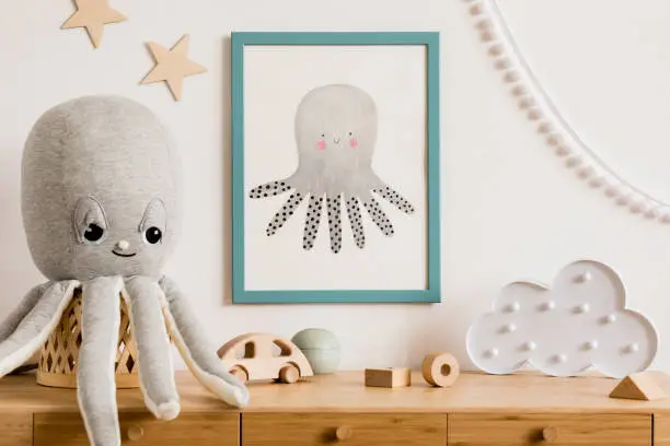Photo of Stylish and cozy childroom with white mock up photo frame, wooden accessories, toys, clouds, plush octopus and white garland and stars on the wall. Bright and sunny interior. Template, Real photo.