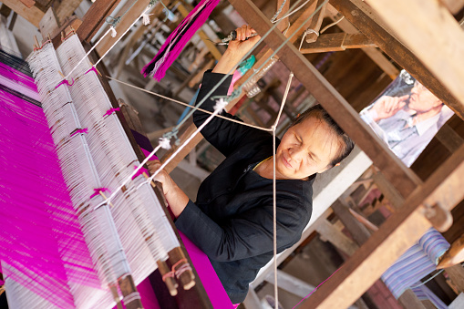 Thai loom craft works scene with thai senior woman in Sun-Phan Tong. City for craft works in Chiang Mai province. Above woman is a picture of King Bhumipol at loom. Outdoor shot of woman working under attached roof. No factory area, local street working woman in Sun-Phan Tong. Showing craft works is part of tourism in city.