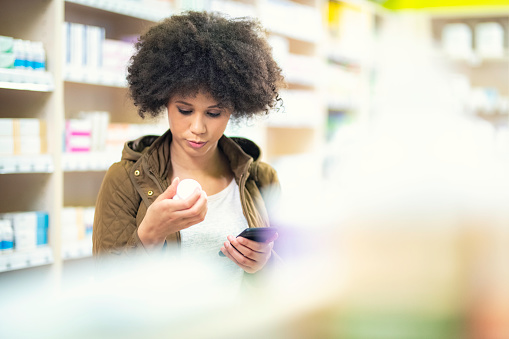 Young woman holding smartphone and reading label of medicine bottle in drugstore.