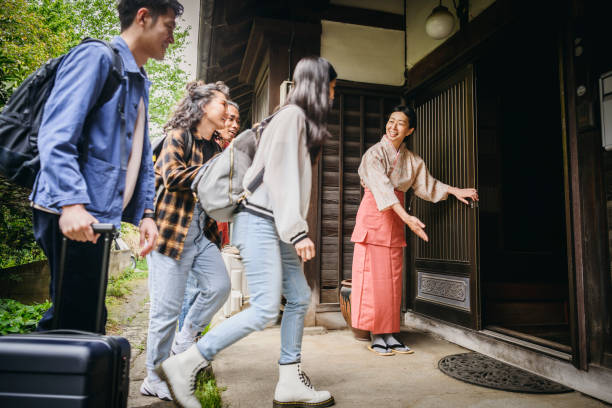 Group of young travellers arriving at traditional Japanese ryokan inn Japanese women welcoming four backpackers with luggage, opening door, ushering them inside the building bed and breakfast stock pictures, royalty-free photos & images