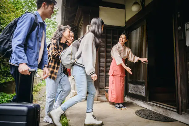 Japanese women welcoming four backpackers with luggage, opening door, ushering them inside the building