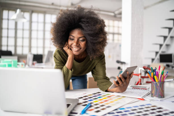 African Fashion designer working in studio and using laptop and smart phone Fashion designer working in studio. Young black woman in an office smiling to camera, close up fashion designer stock pictures, royalty-free photos & images