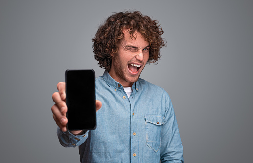 Handsome guy in blue shirt winking and looking at camera while standing on gray background and showing modern smartphone with empty display