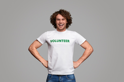 Attractive male in volunteer T-shirt cheerfully smiling and keeping fists on waist while standing at gray background and looking at camera
