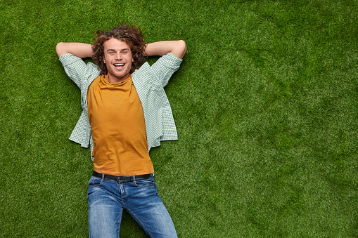 From above cheerful young guy in casual outfit cheerfully smiling and keeping hands behind head while resting on green lawn near copy space