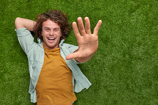 Cheerful young guy with curly hair looking at camera and trying to reach out camera with hand while resting on green lawn