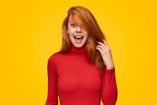 Lovely young woman in red turtleneck sweater touching ginger hair and looking at camera with amazed face expression while standing on vibrant yellow background