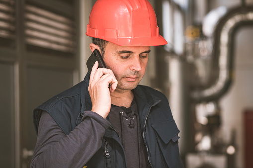 Male Worker with Protective Helmet is Standing in the Heating Plant and Talking over his Mobile Phone. Heating Engineer is in the Power Station Surrounded by Controlling Equipment.