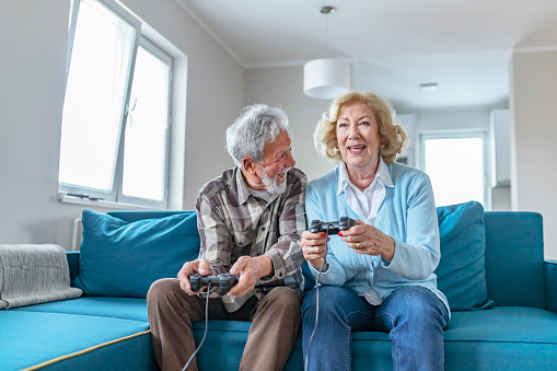 Happy Senior Couple Sitting Together on Their Living Room Sofa Holding Controllers and Laughing While Playing a Video Game Console. Excited Elderly Couple Sitting on a Sofa in Their Living Room in Front of the Television Screen Playing Computer Games